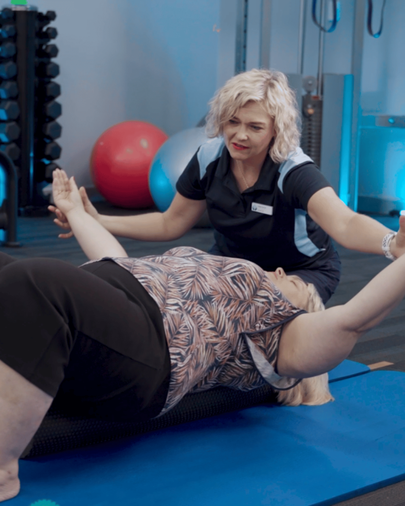 Assist Allied Health physiotherapist guiding a client lying on a massage roller, arms spread out, while providing expert assistance with exercises and strengthening for improved musculoskeletal health.